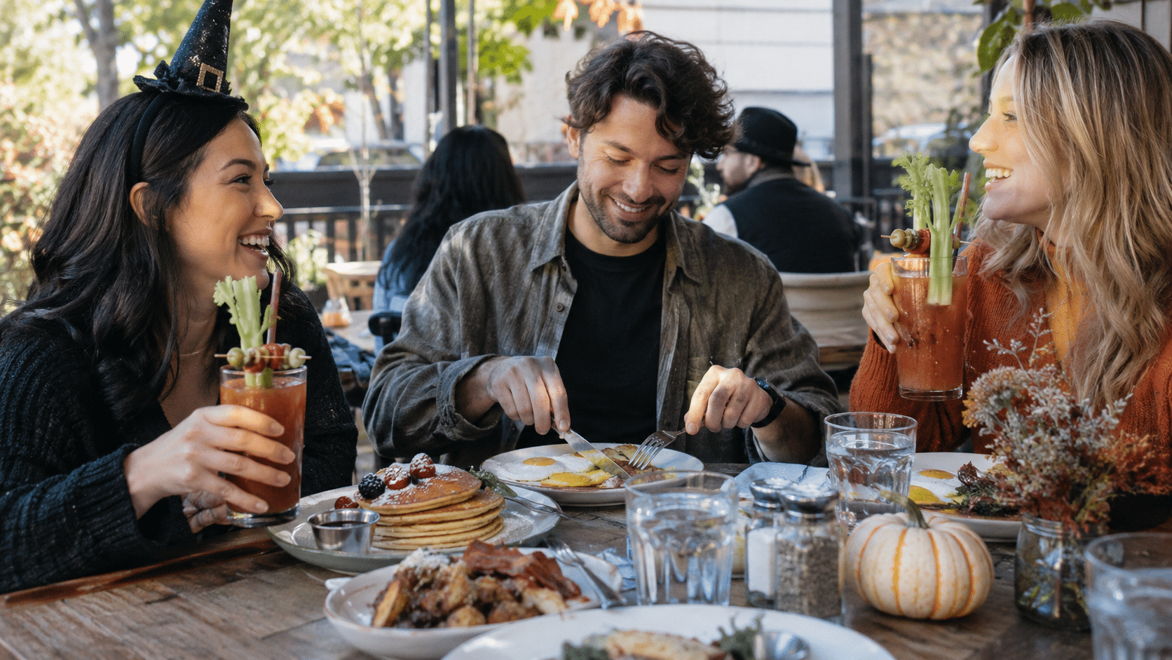 Friends enjoy brunch with pancakes, eggs and garnished tomato drinks beside a small pumpkin.